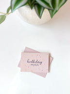 Pink card with 'birthday hugs' text on a white surface with a plant in the corner.