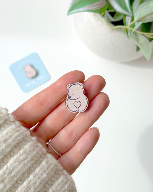 A small bear-shaped wooden token with a heart held on a person's hand. The person is wearing a cream-coloured knit sweater, and a potted plant and a small blue card are blurred in the background.