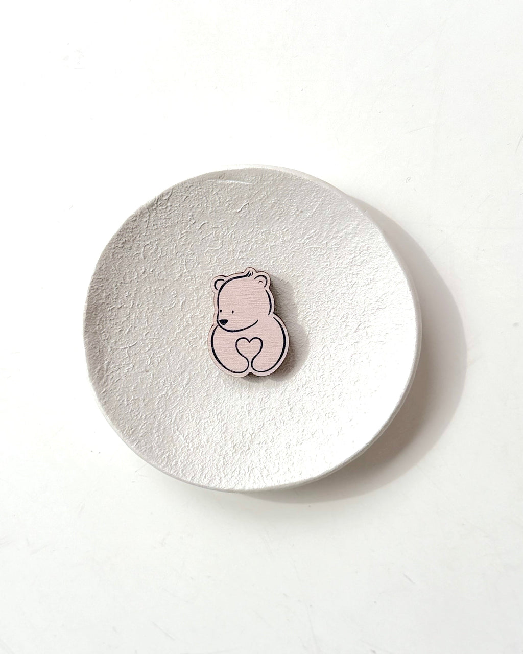 A small pink bear-shaped pin with a heart detail, displayed on a textured round white coaster against a clean white background.