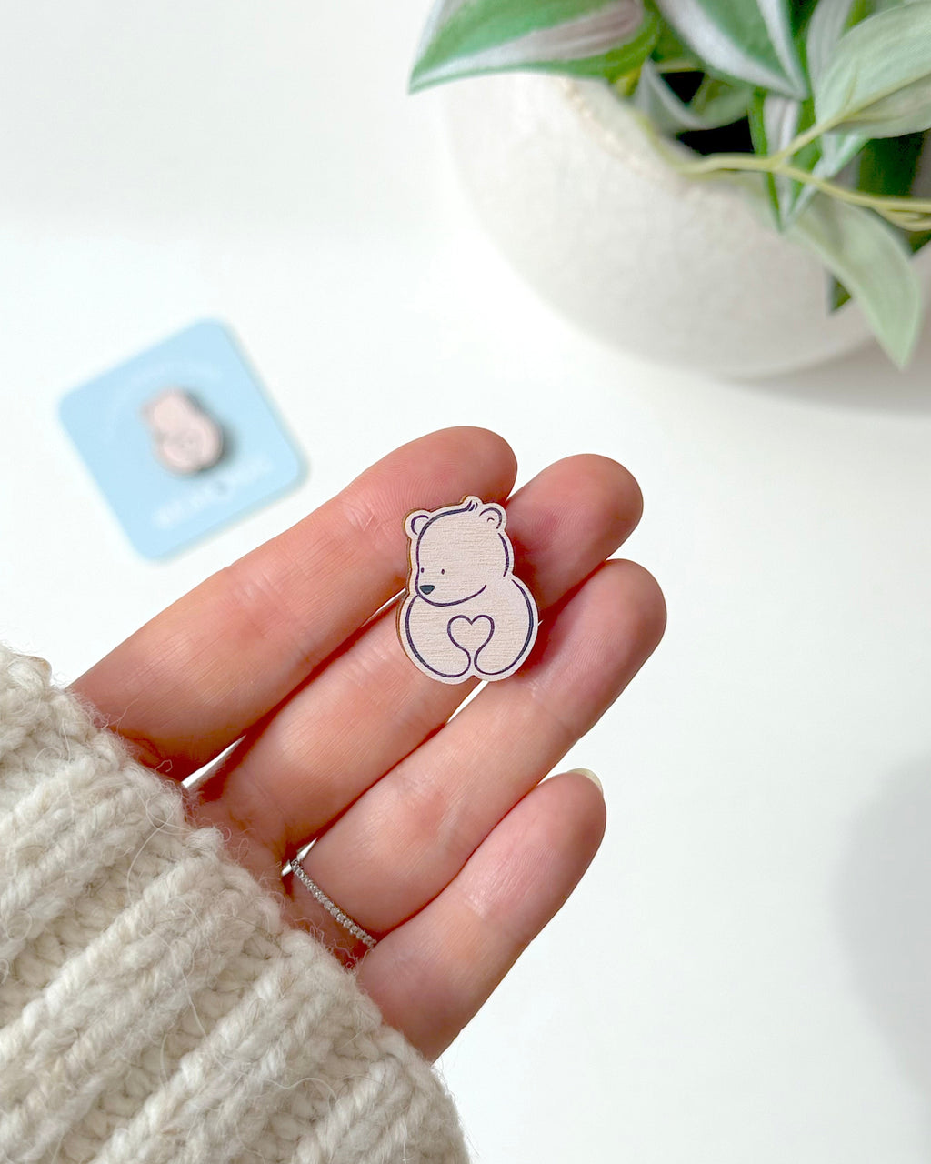 A small bear-shaped wooden token with a heart held on a person's hand. The person is wearing a cream-coloured knit sweater, and a potted plant and a small blue card are blurred in the background.