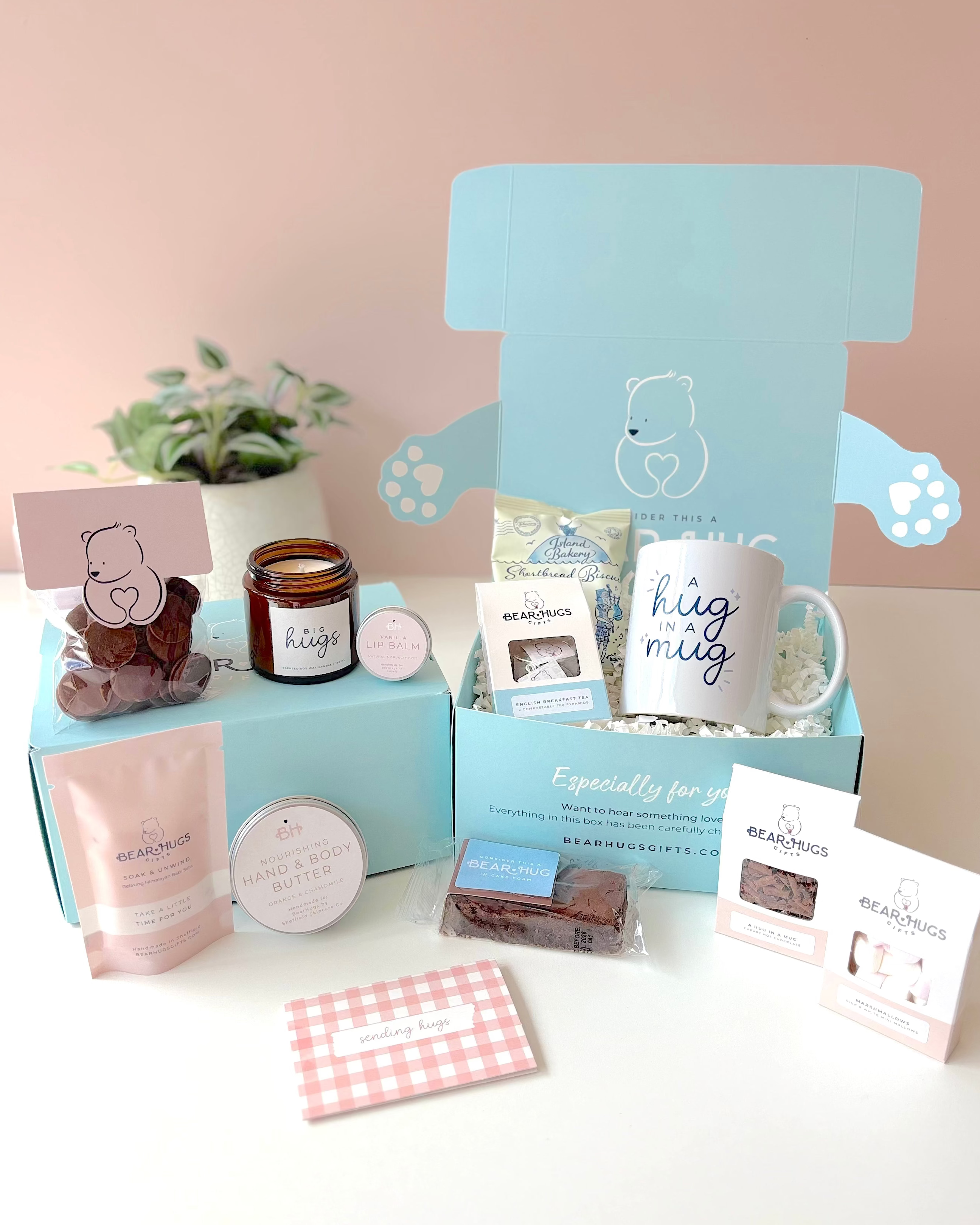 A curated gift box set displayed on a white surface against a soft pink background. The set includes a large light blue gift box with bear-themed cutouts and a mug reading 'hug mug', a smaller blue box with a bear-shaped candle, a pink pouch, a small jar, chocolates, and cards.