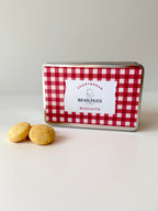 A small rectangular tin of BearHugs Biscuits with a red and white gingham pattern, with two golden-brown biscuits placed in front on a clean white surface.
