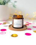 An amber glass jar candle with a lit wick sits on a round wooden tray, surrounded by colorful confetti on a white surface with a blurred potted plant and warm fairy lights in the background. The candle label reads 'Happy Birthday'.