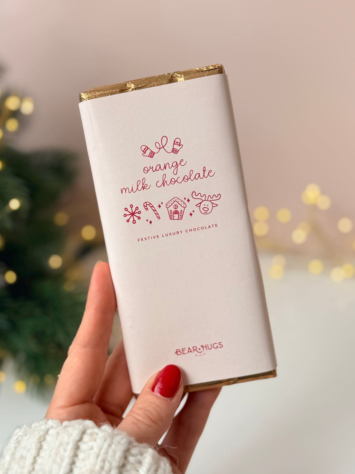 A hand with red nail polish holds a white chocolate bar wrapped in white paper with gold foil edges and pink text reading 'orange milk chocolate' and 'BEARMUGS'. The background is a soft-focus indoor setting with a Christmas tree and warm string lights.