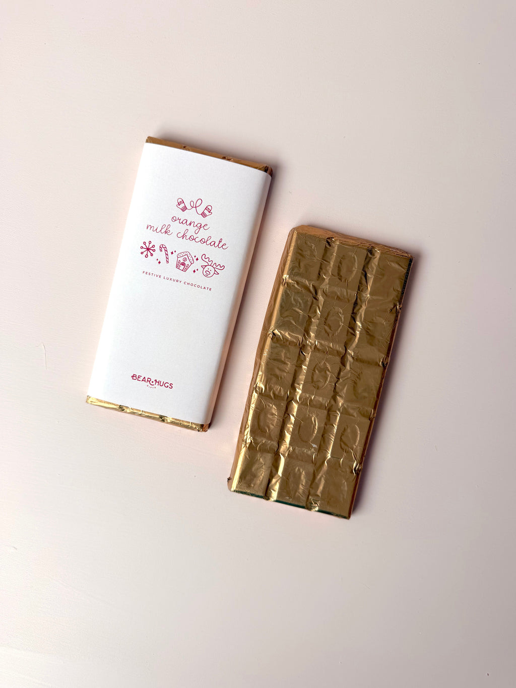 A milk chocolate bar shown next to its white wrapper with pink text and gold trim, alongside the gold foil-wrapped chocolate bar itself, on a light beige background.