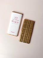 A milk chocolate bar shown next to its white wrapper with pink text and gold trim, alongside the gold foil-wrapped chocolate bar itself, on a light beige background.