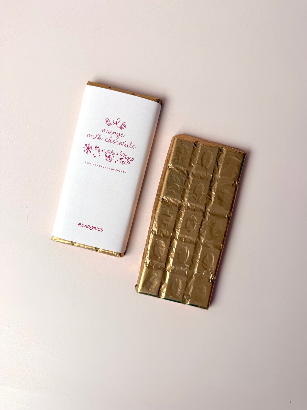 A milk chocolate bar shown next to its white wrapper with pink text and gold trim, alongside the gold foil-wrapped chocolate bar itself, on a light beige background.