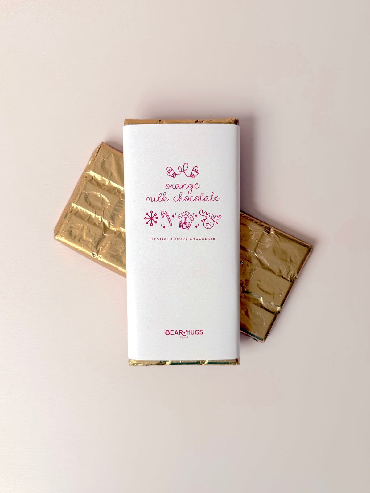 A wrapped milk chocolate bar in the center with two gold chocolate bars behind it on a plain light beige background. The front wrapper is white with pink text reading 'orange milk chocolate' and small floral illustrations, plus the brand name 'earl+mugs' near the bottom.
