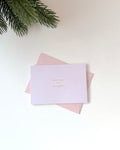 A light lavender greeting card with gold foil text reading 'merry and bright' resting on top of a pale pink envelope, styled on a white surface with pine branches in the top-left corner.