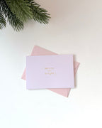 A light lavender greeting card with gold foil text reading 'merry and bright' resting on top of a pale pink envelope, styled on a white surface with pine branches in the top-left corner.