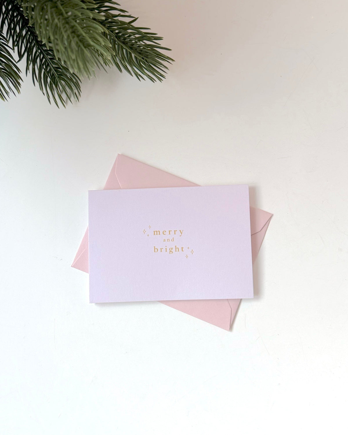 A light lavender greeting card with gold foil text reading 'merry and bright' resting on top of a pale pink envelope, styled on a white surface with pine branches in the top-left corner.
