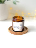 An amber glass jar candle with a lit wick sits on a round wooden coaster. The label reads 'Happy New Home' with a key and heart design.