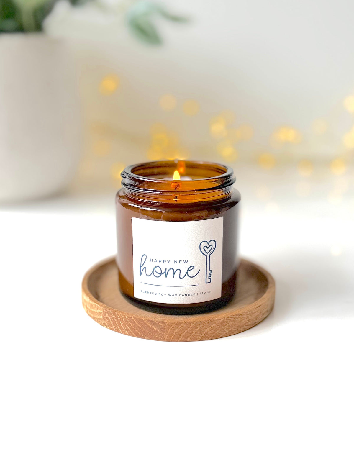 An amber glass jar candle with a lit wick sits on a round wooden coaster. The label reads 'Happy New Home' with a key and heart design.