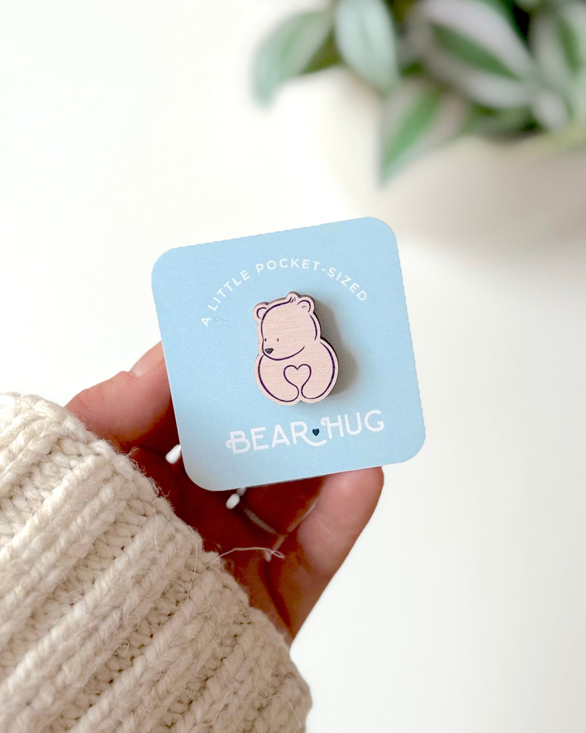 A hand wearing a cream ribbed sweater holds a light blue card displaying a small pink bear hug pocket token with a heart, with a blurred green plant in the background.