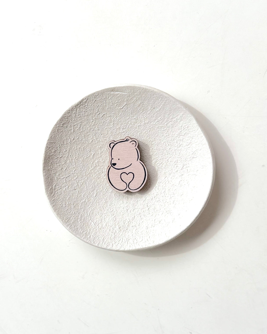A small pink bear-shaped pin with a heart detail, displayed on a textured round white coaster against a clean white background.
