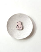 A small pink bear-shaped pin with a heart detail, displayed on a textured round white coaster against a clean white background.