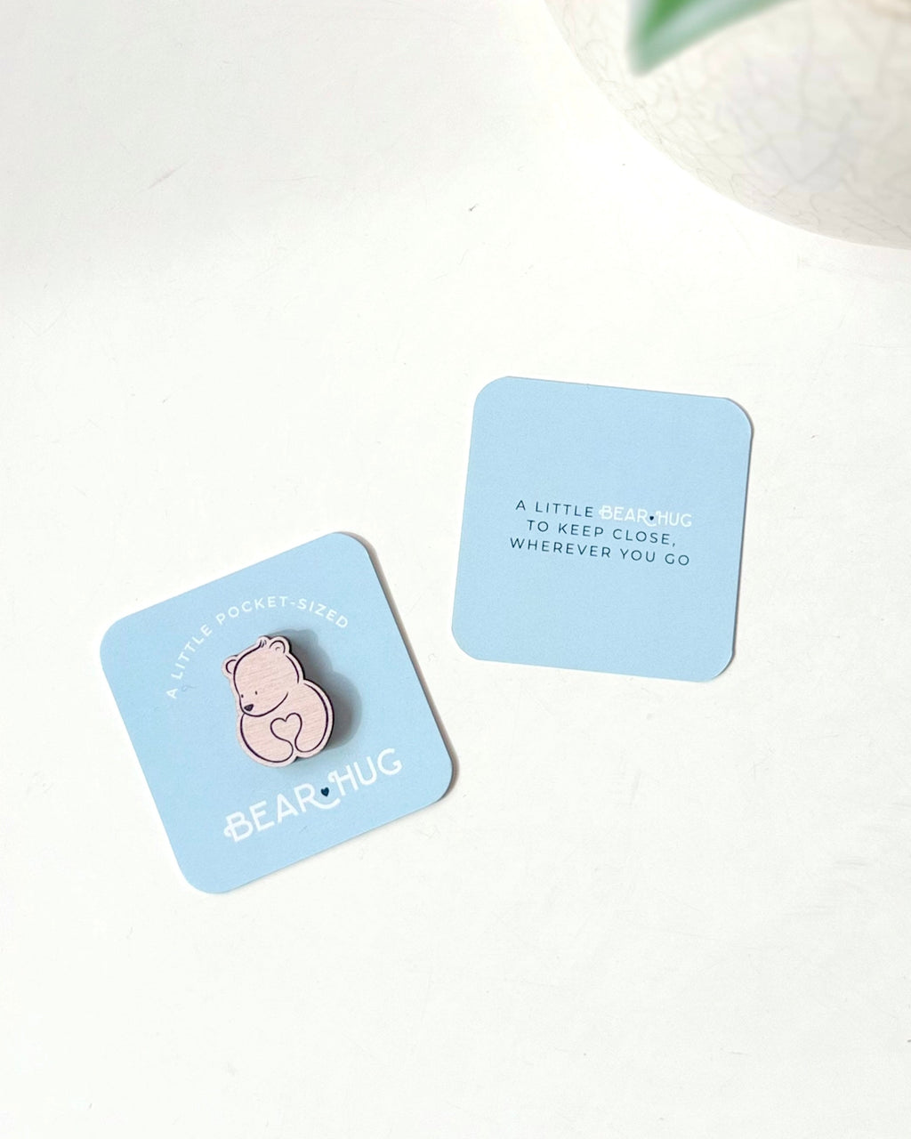 Two light blue square cards on a white surface, one featuring a pink bear-shaped wooden token and the text 'A LITTLE BEAR HUG TO KEEP CLOSE WHEREVER YOU GO' and 'BEARHUG', and the other with the text 'A LITTLE BEAR HUG TO KEEP CLOSE WHENEVER YOU GO'. A white textured planter with green leaves is partially visible in the top right corner.