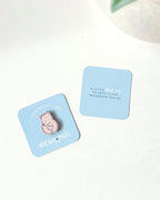 Two light blue square cards on a white surface, one featuring a pink bear-shaped wooden token and the text 'A LITTLE BEAR HUG TO KEEP CLOSE WHEREVER YOU GO' and 'BEARHUG', and the other with the text 'A LITTLE BEAR HUG TO KEEP CLOSE WHENEVER YOU GO'. A white textured planter with green leaves is partially visible in the top right corner.