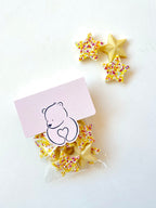 Star-shaped sugar cookies with colorful sprinkles, shown with a small clear bag and a pink card featuring a teddy bear illustration, on a white background.